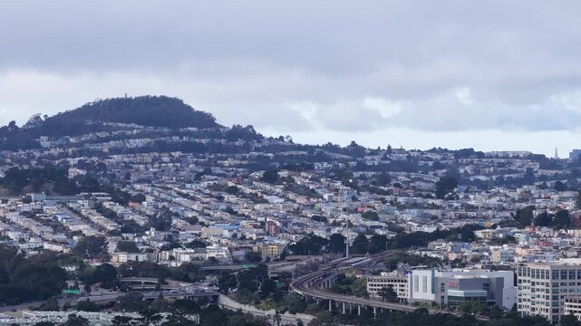 Aerial view showing the sprawling Broadmoor residential neighborhood in Daly City, California, with houses covering the hills under a cloudy sky and the bart railway line running through the city