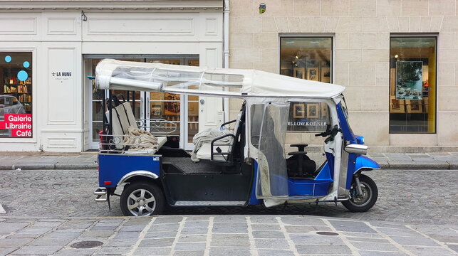A blue electric three-wheeled taxi is parked on a historic cobblestone street alongside upscale retail storefronts featuring large glass display windows in Paris, France
