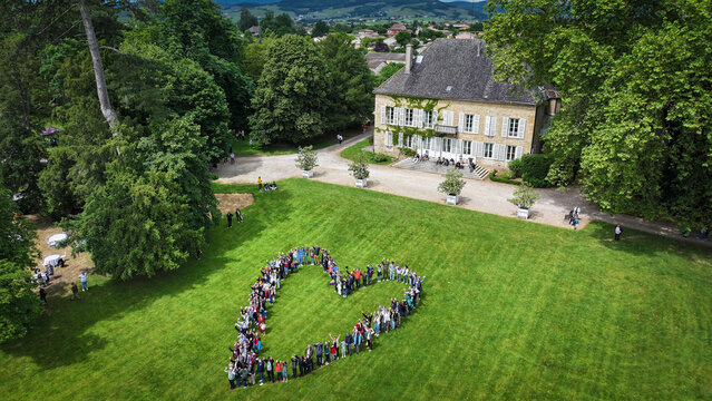 La Chapelle-de-Guinchay, France - 01 June 2024: Aerial view of a large group of people forming a heart shape on a green lawn in front of a historic manor house surrounded by lush trees.