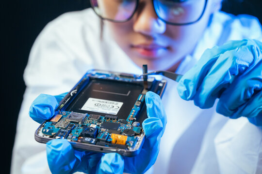 young female electronics technician carefully repairs a smartphone at her workstation. She uses professional precision screwdrivers and tweezers under a bright workspace light. 