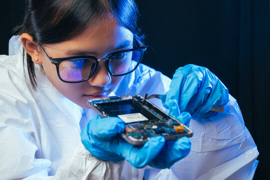 young female electronics technician carefully repairs a smartphone at her workstation. She uses professional precision screwdrivers and tweezers under a bright workspace light. 