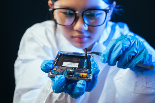 young female electronics technician carefully repairs a smartphone at her workstation. She uses professional precision screwdrivers and tweezers under a bright workspace light. 