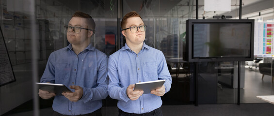 Professional man, corporate staff member with Down syndrome stands next to glass blackboard with digital tablet in office, looks away with pensive expression, thinks on ideas, planning, strategizing