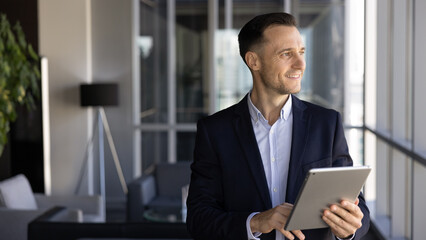 Fototapeta premium Confident and successful businessman stands by office window, smiling, reflecting on creative ideas, reviewing project updates on digital tablet. Strategic thinking, business decisions, technology