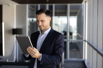 Fototapeta premium Focused businessman stands in office hallway with tablet, managing business tasks, reads information, review digital data, consulting corporate client via email, staying connected through technology