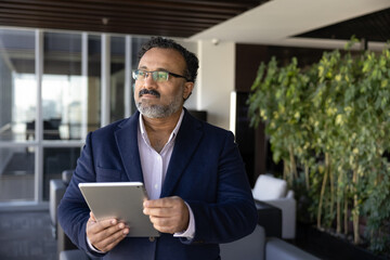 Fototapeta premium Thoughtful middle-age businessman stands in modern office with digital tablet, reviewing information, evaluating received data, thinking on solution or idea, planning strategic steps or making