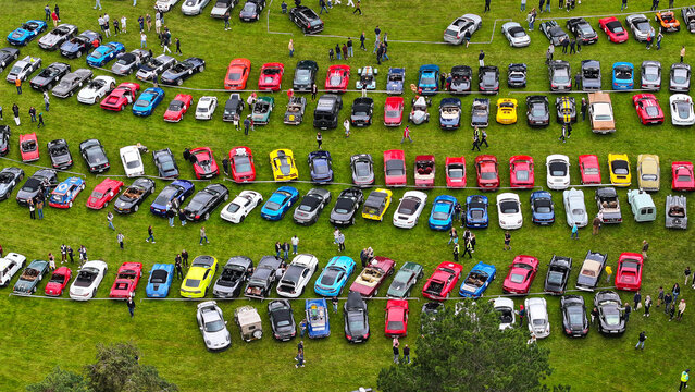 La Chapelle-de-Guinchay, France - 01 June 2024: Aerial view of rows of colorful sports cars and classic vehicles parked on a lush green field with people walking between them.