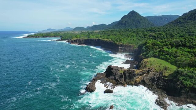 Aerial drone footage of Swadaya camping ground, on the rocky cliff coast nearby Sekepung beach, in south Sumbawa island, Indonesia, with huge forests and mountains behind, rough waves and blue waters