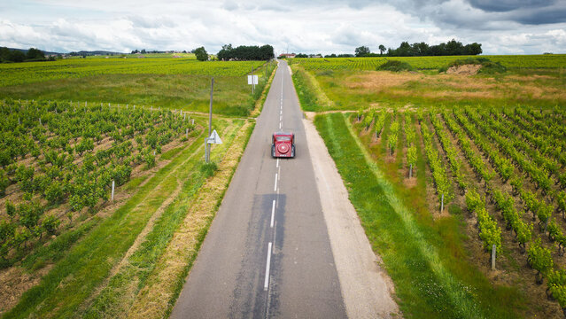 Aerial view of a vintage red car driving on a straight road through lush green vineyards under a cloudy sky La Chapelle-de-Guinchay, Bourgogne-Franche-Comte, France.