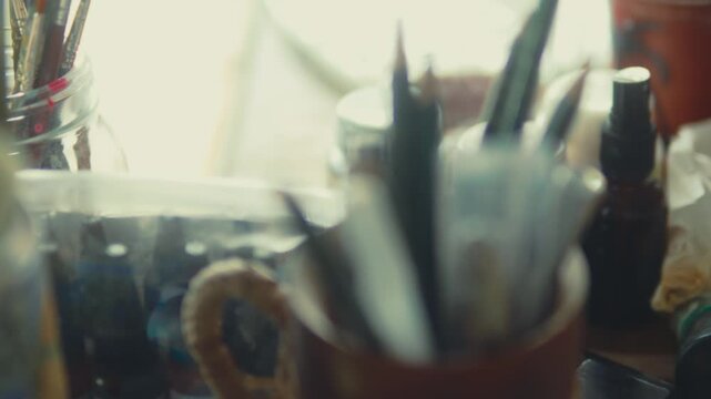 Camera moves across cluttered art table with brushes paint tools and materials