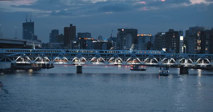 Wide view, train crossing river in blue evening twilight on urban city backdrop. Modern Tokyo cityscape view at dusk. Commuter train crossing an illuminated railway. Tour boats moving on water below
