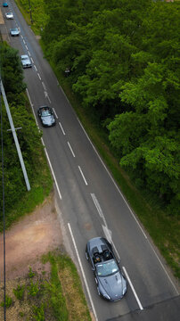 Aerial view of luxury sports cars driving along a paved road bordered by dense green trees and foliage in La Chapelle-de-Guinchay, Bourgogne-Franche-Comte, France.