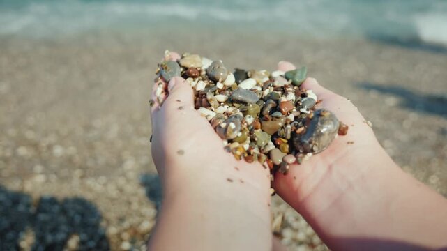 Hands holding small wet sea pebbles on sunny beach background with waves motion.