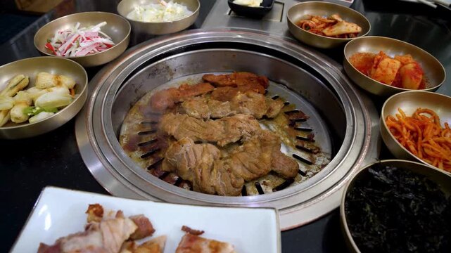 Korean barbecue grill with sizzling marinated pork belly surrounded by traditional banchan side dishes in brass bowls during authentic dining experience.