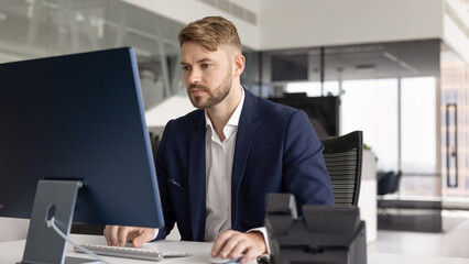 Fototapeta premium Young man, office employee sits at desk, read emails or important correspondence, writing or editing proposal and document, evaluating financial or project planning information, strategizing using PC
