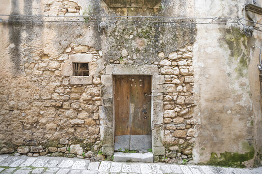 View of a rustic wooden door set in an ancient stone wall with a small barred window and mossy textures in Ragusa, Sicily, Italy.