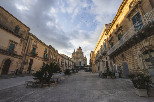 View of the Duomo di San Giorgio at the end of a historic street lined with baroque architecture and palm trees under a cloudy sky in Ragusa, Sicily, Italy.