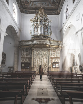 View of the ornate golden pipe organ and wooden pews inside a historic church with a woman standing in the aisle in Polignano a Mare, Apulia, Italy.