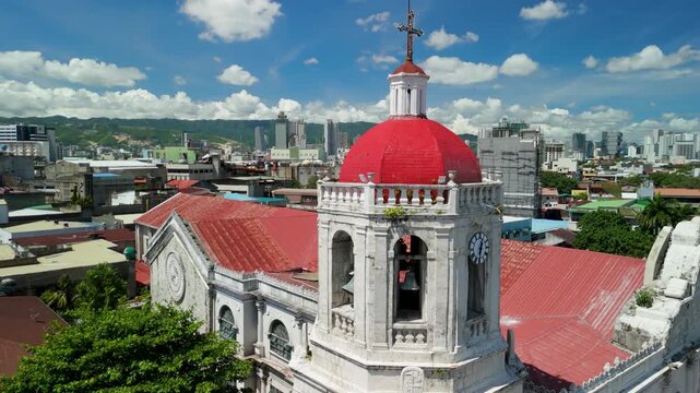 Aerial parallax around Cebu Metropolitan Cathedral's ornate bell tower and iconic red dome, with clock, bronze bells, cross, and Cebu City skyline and mountains in the background