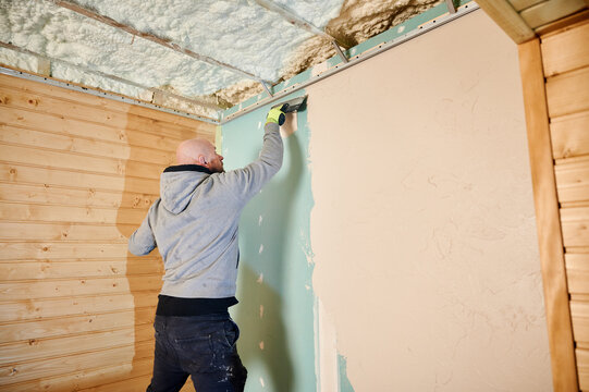 Man applying wall putty with trowel, smoothing drywall surface during interior renovation. Worker leveling plaster layer near ceiling with insulation visible, home finishing and construction concept