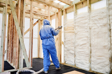 Fototapeta premium Worker spraying polyurethane foam insulation into timber wall cavities inside unfinished house. Interior construction scene, thermal sealing, weatherization and energy efficient building concept