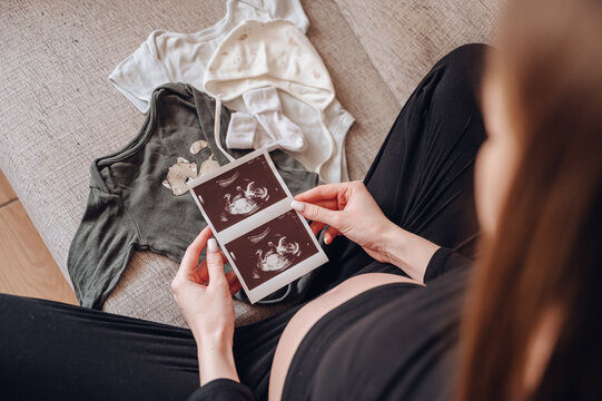 Close up of a pregnant woman holding ultrasound images of her baby next to tiny clothes on a sofa