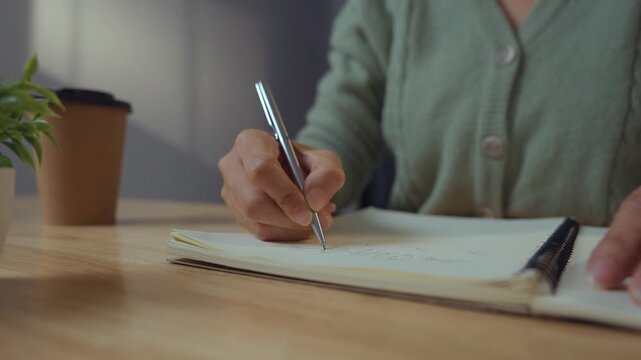 Close-up image of a woman's hand holding a pen and writing on a surface, possibly a notebook or a desk.