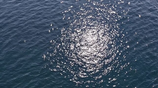 Sunlight reflecting on rippling ocean water surface, creating a shimmering circular pattern, captured from above, showcasing the dynamic movement of waves and light interplay