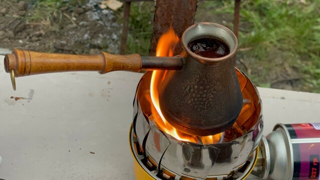 Brewing coffee in a copper cezve over an open flame on a portable gas stove outdoors.