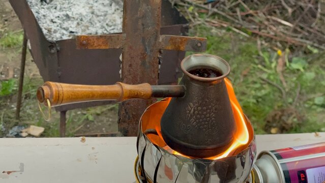 Brewing coffee in a copper cezve over an open flame on a portable gas stove outdoors.