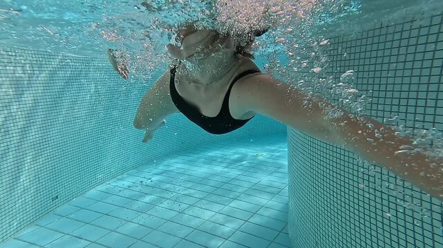 an underwater video shot of a woman in a black swimsuit swimming breaststroke against a counter current in an indoor tiled swimming pool concept of active recreation, water sports, and training