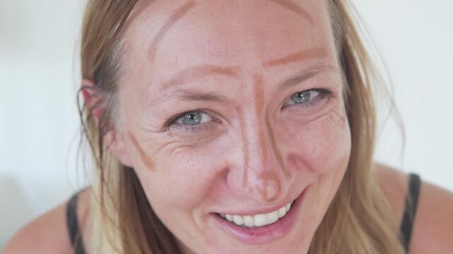 Smiling woman with playful makeup contour lines drawn on her face during a family beauty routine at home. Authentic close-up portrait showing natural skin texture, real emotions, self-care, humor and