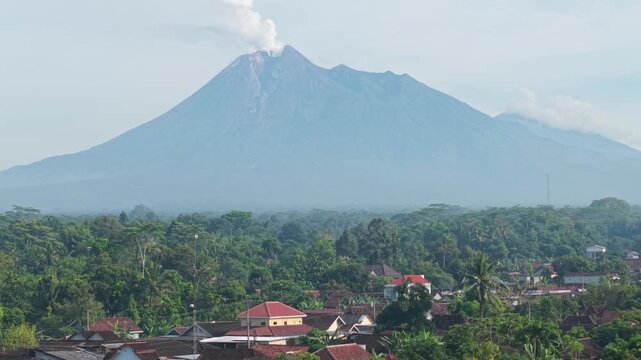 Aerial view of Mount Merapi volcano with smoke rising above lush forests and village rooftops in Kemalang, Central Java, Indonesia.