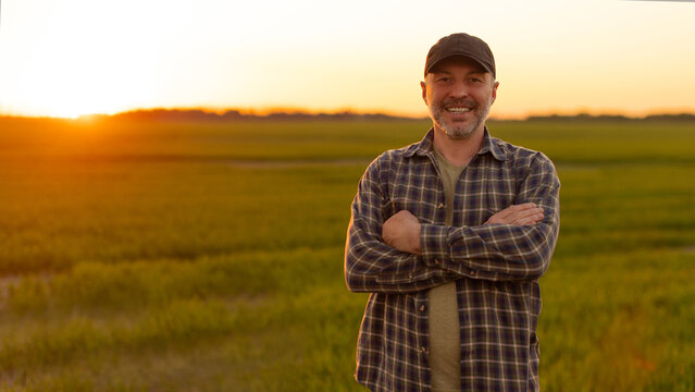 Farmer in cap standing arms crossed in green wheat field at sunset