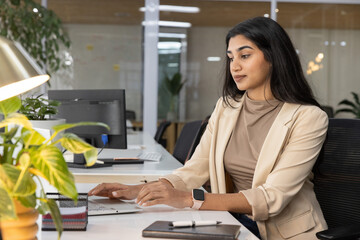 Indian woman wearing beige blazer typing on laptop at open-plan office desk checking smartwatch