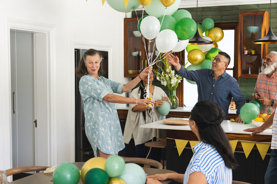 Diverse family and friends arranging balloons in home kitchen, host wearing blue dress, copy space