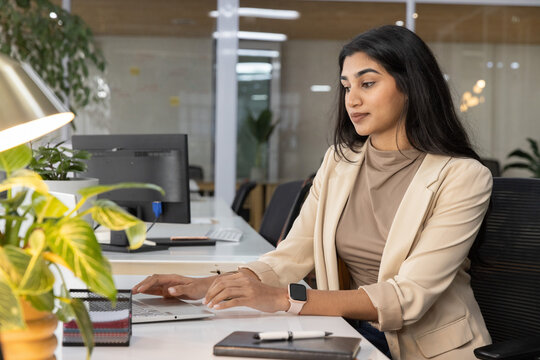 Indian woman wearing beige blazer typing on laptop at open-plan office desk checking smartwatch
