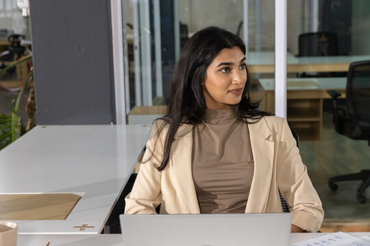 Indian woman sitting at office desk wearing beige blazer using laptop, document, mug, pad