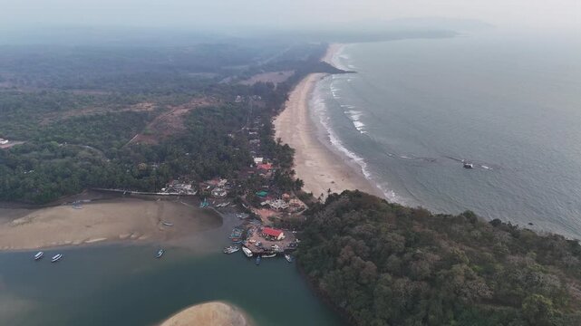 Drone shot capturing a sprawling seaside resort with symmetrical red-roofed buildings, landscaped lawns, and a central pool area, positioned between river channels and a tranquil beach coastline.