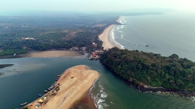 Cinematic aerial view of The LaLiT Golf and Spa Resort Goa, nestled between lush greenery, river backwaters, and the Arabian Sea, with waves rolling along the coastline under a soft hazy sky.