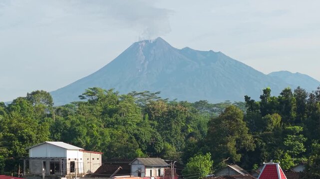 Aerial view of Mount Merapi volcano with smoke rising behind a traditional red tiled roof in Kemalang, Central Java, Indonesia.