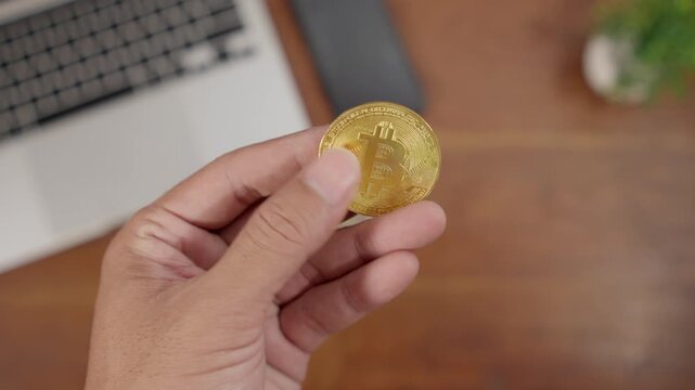 Close-up of a hand holding a gold Bitcoin coin on a work desk, illustrating cryptocurrency, digital finance, blockchain technology, and modern investment concepts.