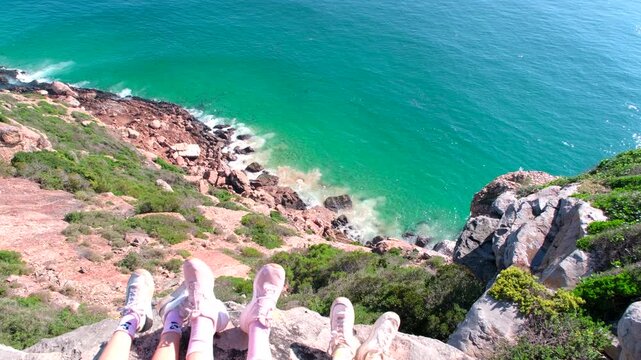 View over hikers' feet dangling off boulder down sea cliff at Robberg reserve
