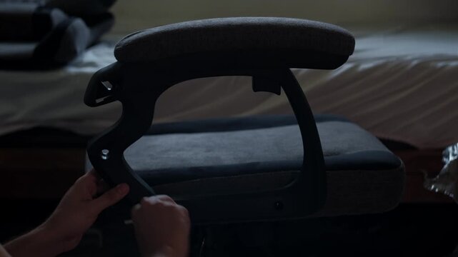 Medium shot of hands installing a gaming chair armrest indoors during furniture assembly with dim lighting and visible tool use