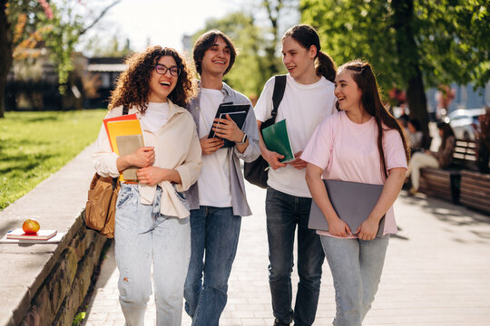 Group of four diverse students walking together on a sunny day, carrying books and tablets, with green grass and trees in the background
