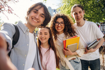 Group of four diverse students smiling for a selfie outdoors, holding books and notebooks, surrounded by greenery and urban architecture in the background © standret