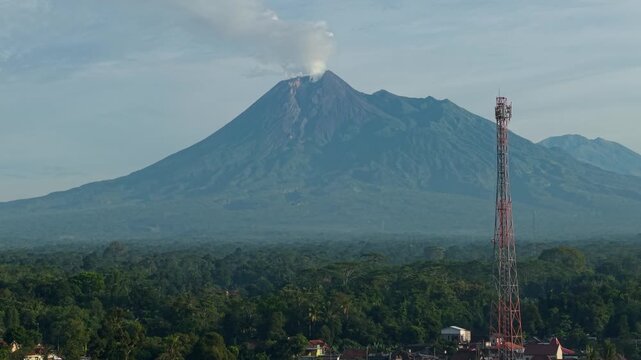 Aerial view of Mount Merapi volcano emitting smoke above a lush tropical forest and a telecommunications tower in Kemalang, Central Java, Indonesia.