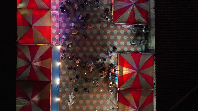 Aerial view of GOR Gelarsena Klaten Night Market with red and white striped tents and people on patterned tiles at night in Kecamatan Klaten Utara, Jawa Tengah, Indonesia.