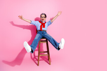 Obraz premium Joyful senior woman jumping from a wooden stool against a pink background, celebrating freedom, confidence, playful style and positive aging in casual denim and white sneakers