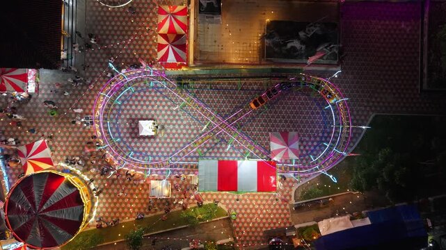 Aerial view of GOR Gelarsena Klaten Night Market featuring a neon-lit roller coaster track and striped tents at night Kecamatan Klaten Utara, Jawa Tengah, Indonesia.